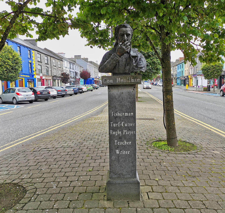 Bust statue of Con Houlihan on a quiet, colourful street with trees and parked cars.