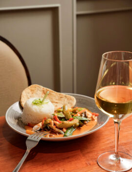 Plate of curry with rice and naan, next to a glass of white wine on a wooden table.