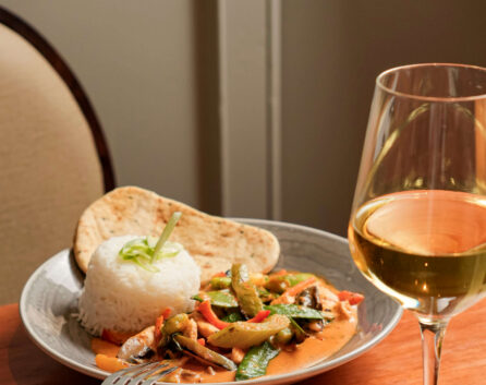 Plate of curry with rice and naan, next to a glass of white wine on a wooden table.