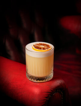 A cocktail with a citrus garnish on a plush red leather chair in a cosy hotel lounge.