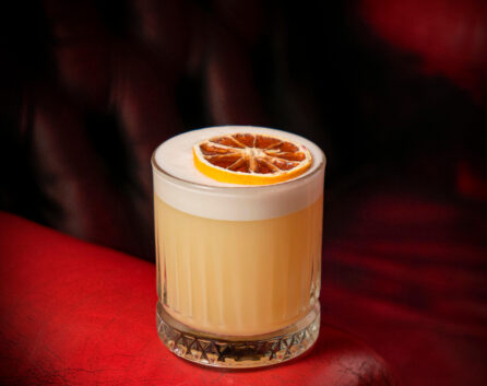 A cocktail with a citrus garnish on a plush red leather chair in a cosy hotel lounge.