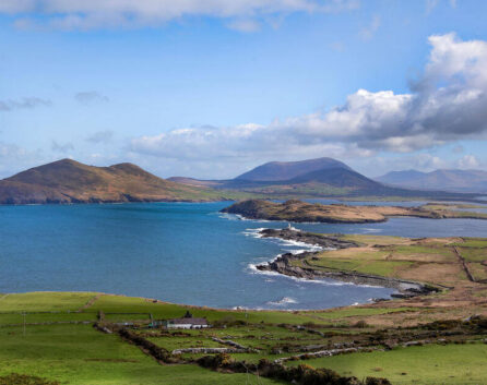 Scenic view of Ring of Kerry coastline, lush fields, and distant mountains under blue sky.
