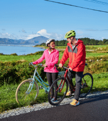 Couple cycling by lush green fields near River Island Hotel with mountains in the background.