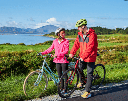 Couple cycling by lush green fields near River Island Hotel with mountains in the background.