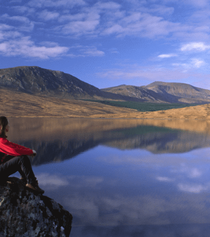 Person in red sitting on a rock by a serene lake with mountains in the background.