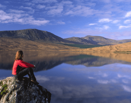 Person in red sitting on a rock by a serene lake with mountains in the background.