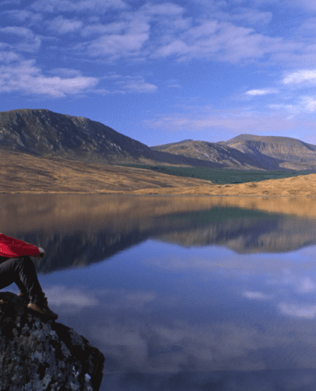 Person in red sitting on a rock by a serene lake with mountains in the background.