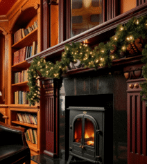 Cosy hotel lounge with festive garland, warm fire, and bookshelves, creating an inviting atmosphere.